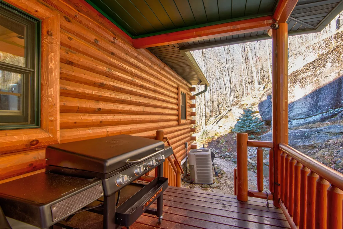 A rustic log cabin deck featuring a grill, wooden railing, and natural landscape views beyond. The grill is ready for barbecues, and the deck offers a seamless transition between indoor comfort and the fresh mountain air.