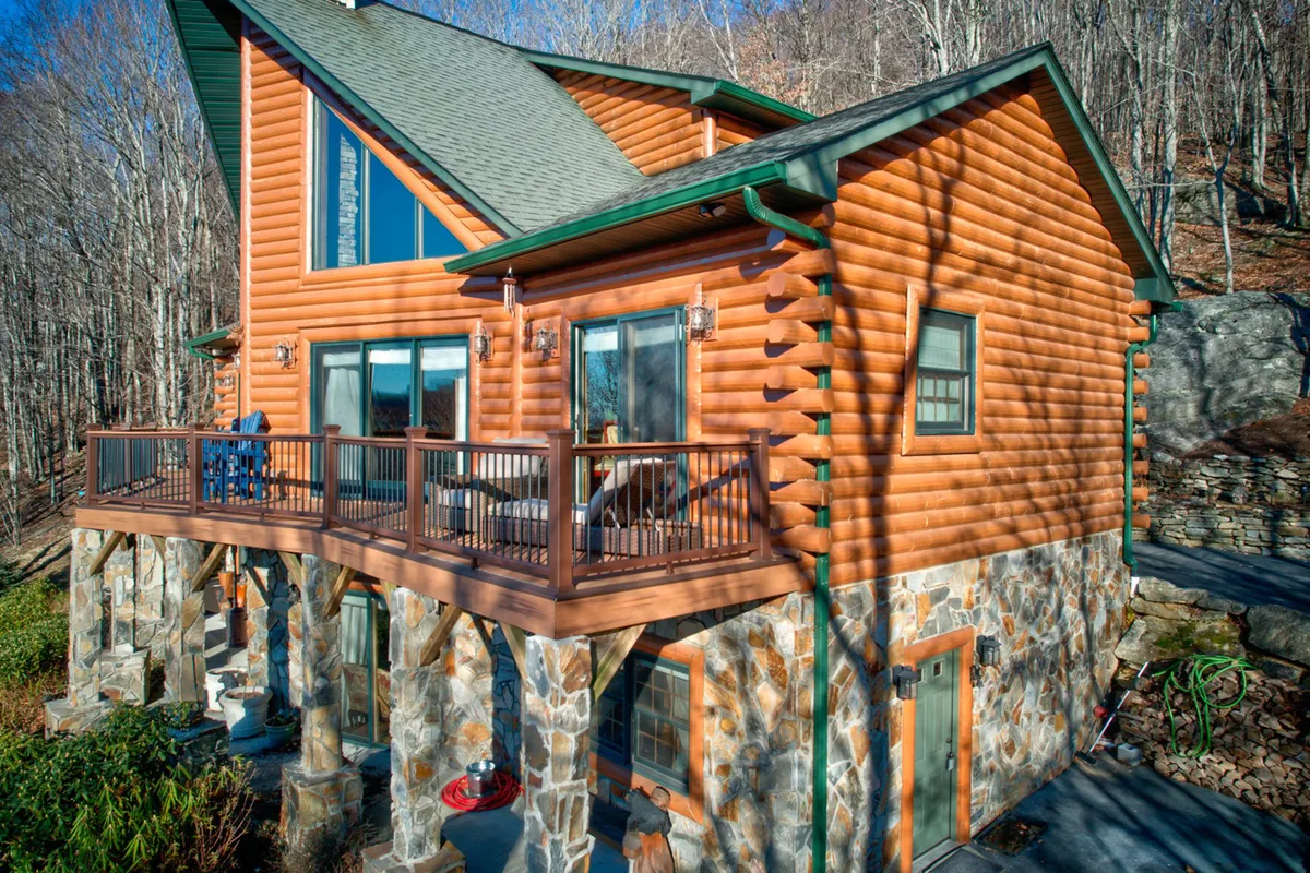 This exterior shot captures the log cabin’s striking architecture, stone foundation, broad windows, and deck area. The cabin is nestled in greenery, emphasizing both its seclusion and proximity to unspoiled nature.