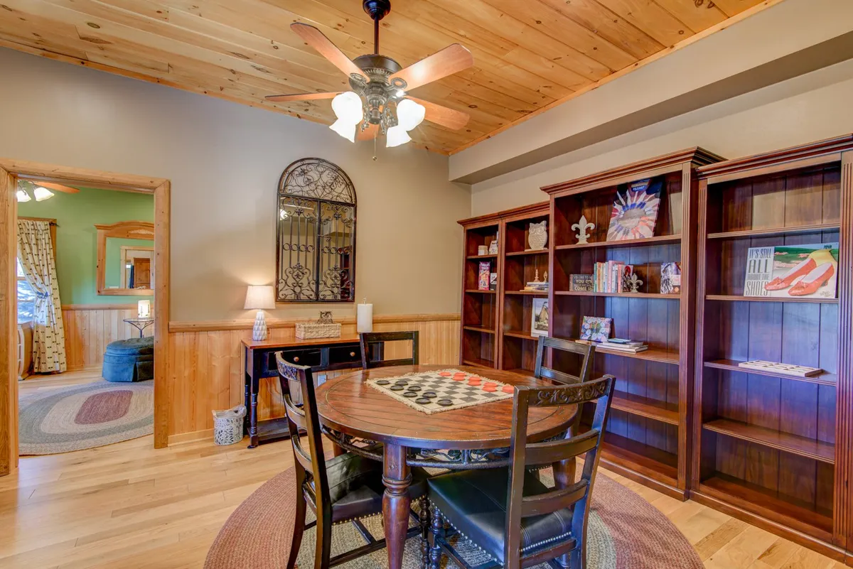 A comfortable interior dining area featuring a table with a built-in checkerboard, surrounded by bookshelves filled with games and books. The space exudes a homey, playful feel perfect for family game nights or casual gatherings. Warm lighting and a ceiling fan complete the cozy, inviting atmosphere.