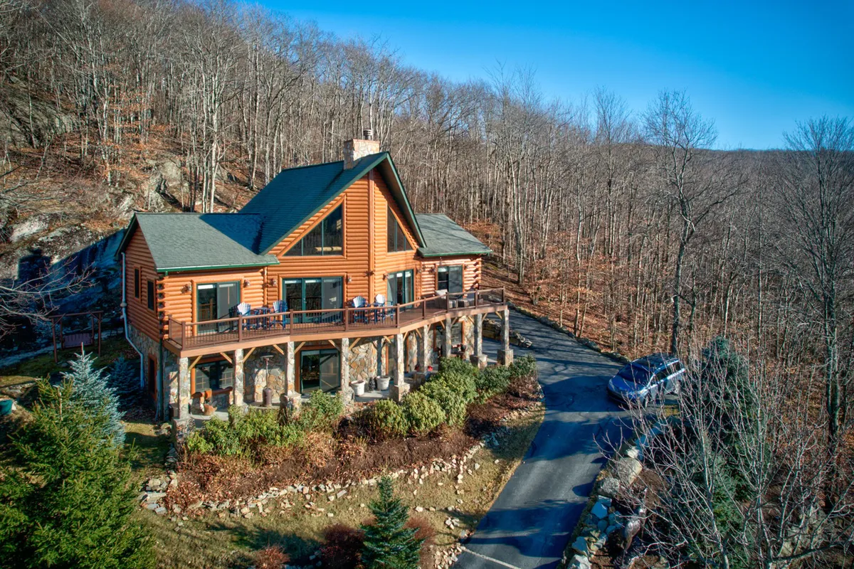 A warm, inviting log cabin with a large deck set among towering trees. The stone foundation adds rustic charm, while the expansive deck is perfect for gatherings. The outdoor setting highlights opportunities for morning coffee, stargazing, and enjoying nature.