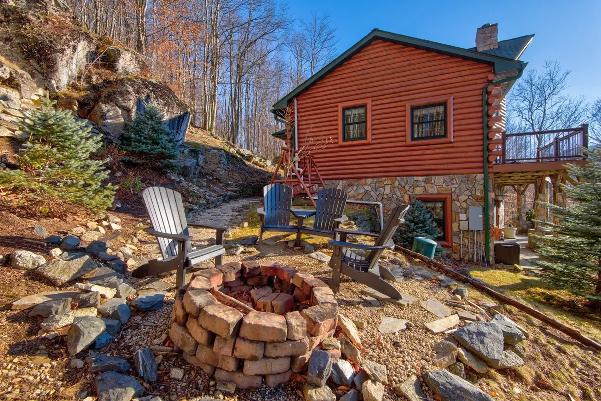 An inviting exterior fire pit area with Adirondack chairs arranged around a crackling fire, set on a stone pathway beside the wooden house and surrounded by trees. The scene radiates warmth and encourages outdoor gatherings for s'mores, stargazing, and storytelling.