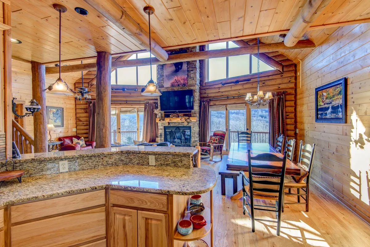 A stunning open-concept great room featuring soaring log beams, a dramatic stone fireplace as the centerpiece, and a wall of expansive windows that frame the mountain scenery. The space includes a large wooden dining table and a granite-topped kitchen island, all bathed in warm, welcoming light. Family members are seen gathering, cooking, and relaxing by the fire, highlighting the lodge’s communal spirit.