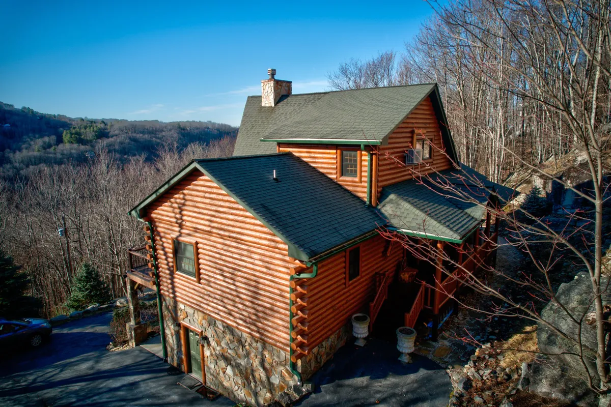 A charming log cabin set on a stone foundation, surrounded by lush trees and distant mountain views. The porch invites relaxation and hints at the seclusion and natural beauty of the area, while the overall scene evokes a sense of adventure and tranquility.