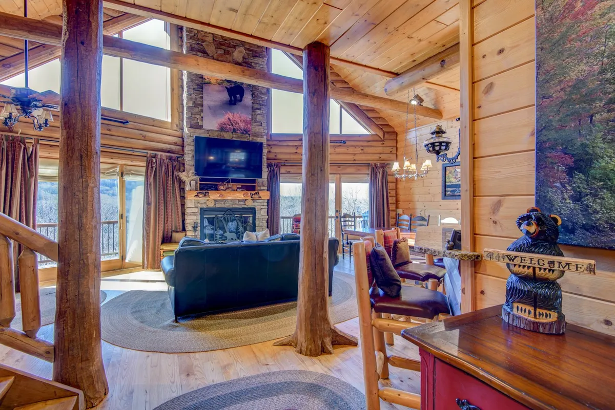 A cozy, inviting interior with a comfortable seating area arranged around a large stone fireplace. The space features a dining table and is set against a wall of windows that allow natural light and mountain views to flood in. The details, like a decorative bear figure and rustic decor, enhance the welcoming, homey feel.