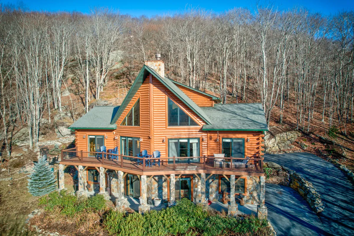 The image showcases a beautiful log cabin nestled among towering trees atop Beech Mountain, with a spacious wraparound deck perfect for gatherings. The warm glow from inside the cabin and the expansive deck invite relaxation, while the backdrop hints at sweeping sunset views.